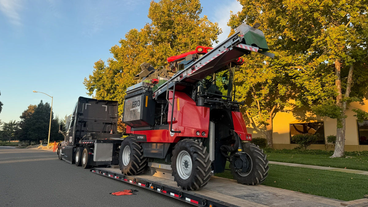 A red and black ERO Grapeliner Harvester with a transfer belt loaded onto a flatbed truck on an outdoor street, with trees and a building in the background.